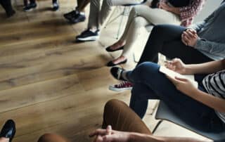 People sitting in a circle on chairs indoors, only their legs and hands are visible on a wooden floor—perhaps a group of mates getting started with an activity together.