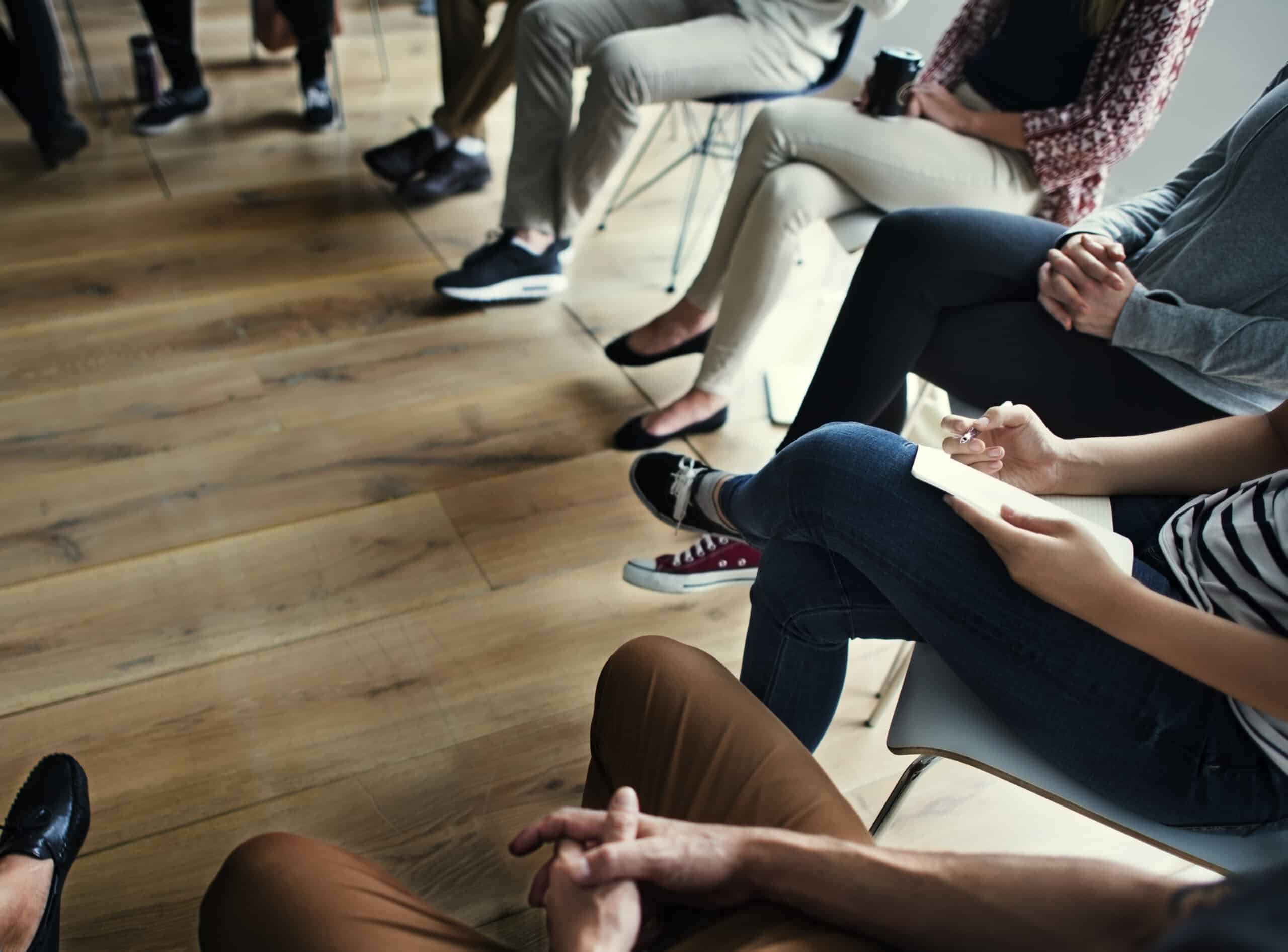 People sitting in a circle on chairs indoors, only their legs and hands are visible on a wooden floor—perhaps a group of mates getting started with an activity together.