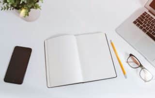 An open notebook for Reading Week study tips sits on a desk with a laptop, smartphone, glasses, pencil, and a plant.